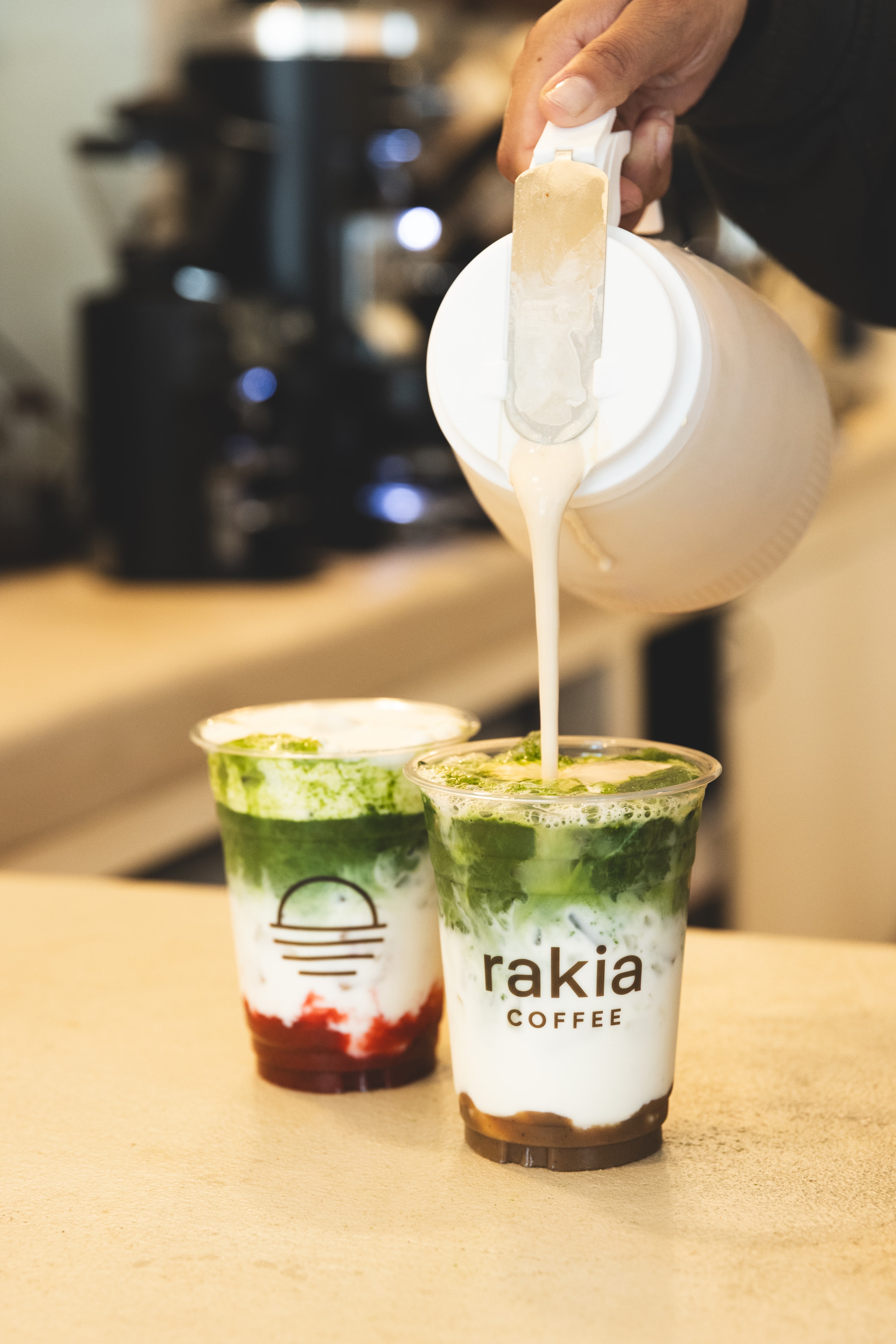 Milk being poured into a layered iced matcha drink at Rakia Coffee in Jacksonville, with a strawberry matcha in the background.